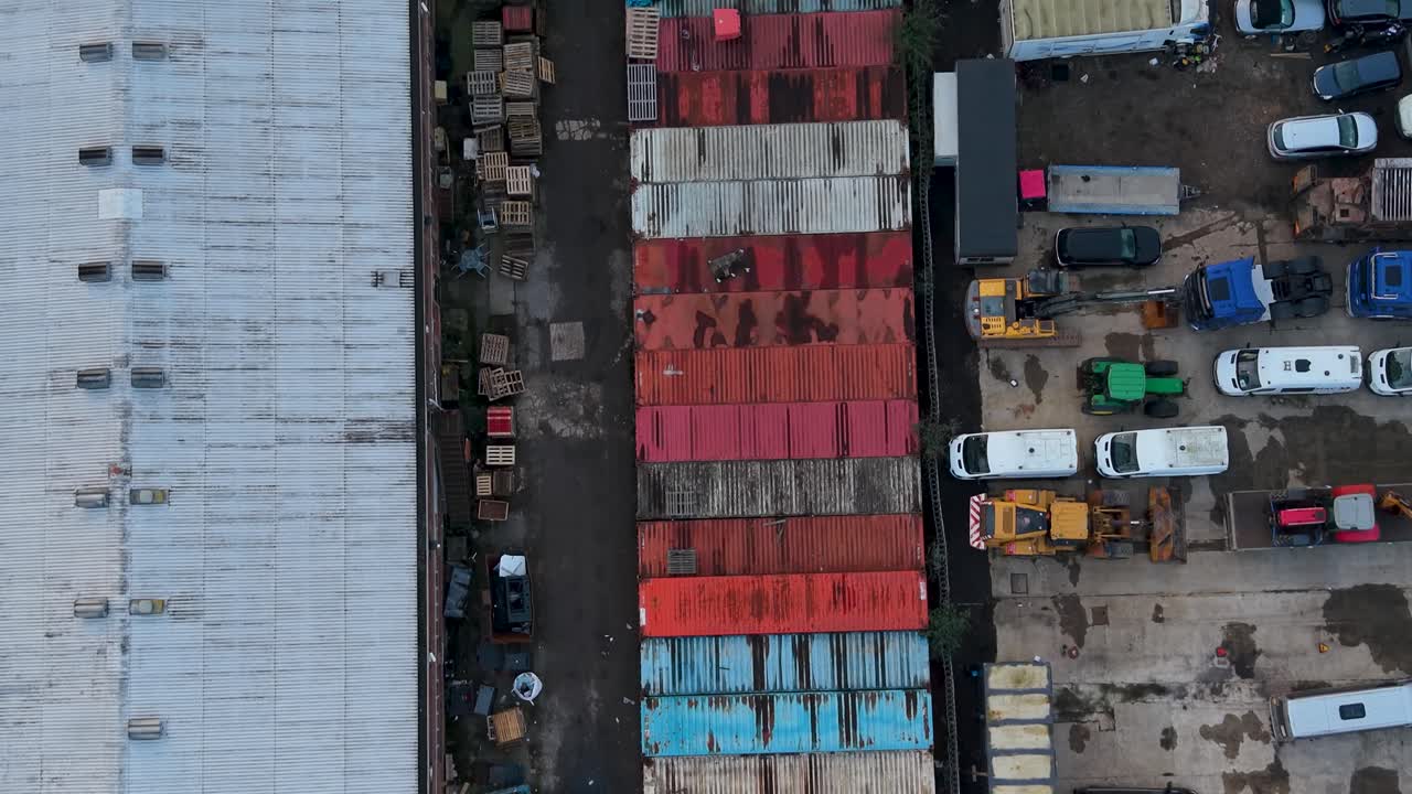 Droneflyvning over fragtcontainere på et industriområde, fremadgående bevægelse set oppefra forbi farverige containerrækker med en skrotplads og parkerede køretøjer ved siden af varehuse.