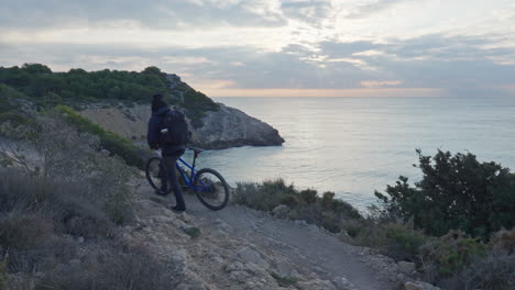 a traveler embarks on a peaceful journey, walking alongside their bike on a pebble trail along the edge of a cliff, as the calm ocean washes in below and the morning sun rises above the horizon