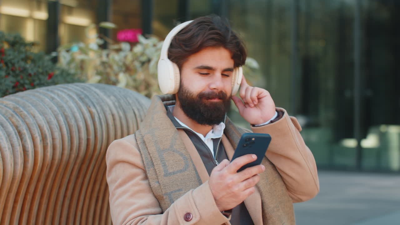 Happy indian man in wireless headphones listening music in smartphone dancing sitting on bench