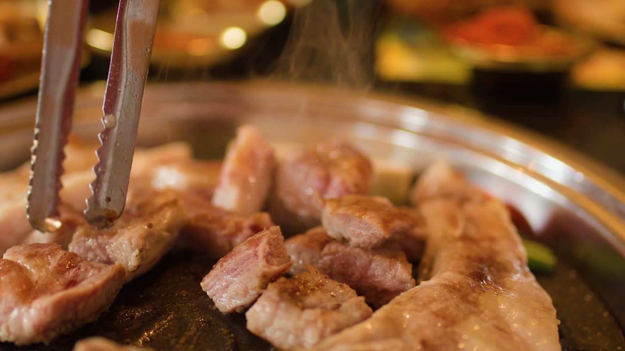Tongs turning pork belly slices on smoky tabletop grill, warm lighting, shallow depth of field