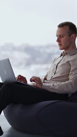 Busy young adult man works on laptop sitting in a chair. Comfortable working atmosphere in spacious room with panoramic windows. Vertical video