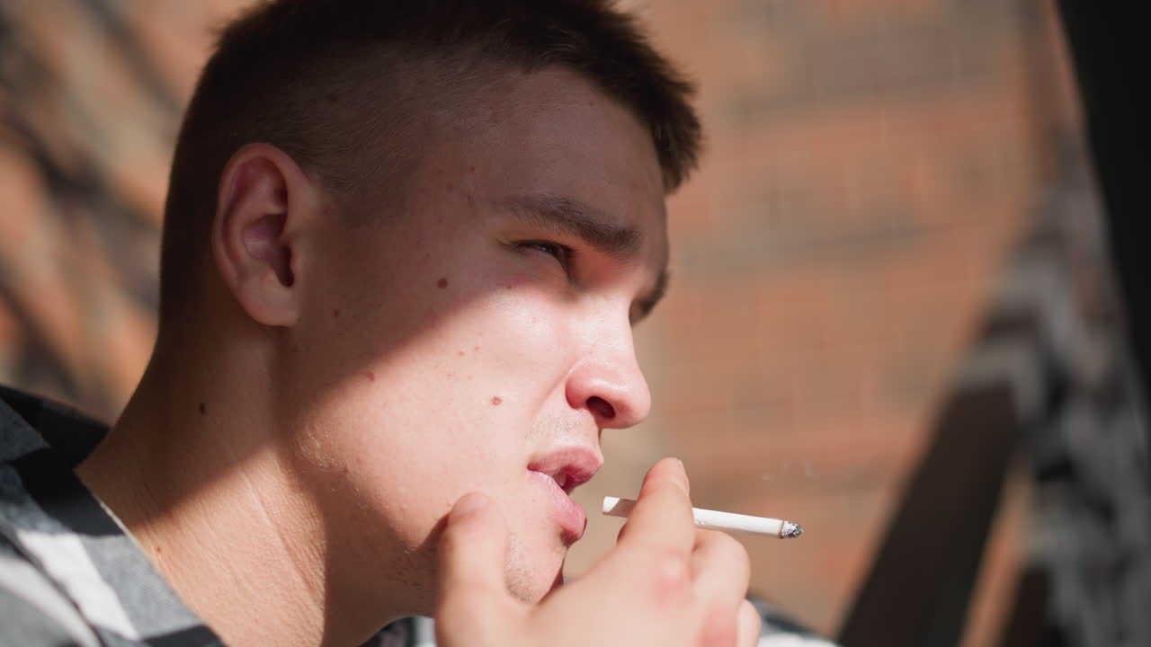 close up of young man with spot on face holding cigarette near lips while smoking thoughtfully against soft blur background with warm sunlight casting shadow across expression beside iron railing outdoors