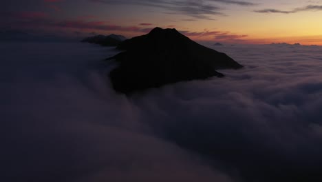 Un-Dron-Inclinado-Hacia-Arriba-De-Un-Hombre-Parado-Solo-En-El-Borde-De-Una-Montaña-Con-Vistas-A-Un-Lecho-De-Nubes-En-Una-Altitud-Extrema