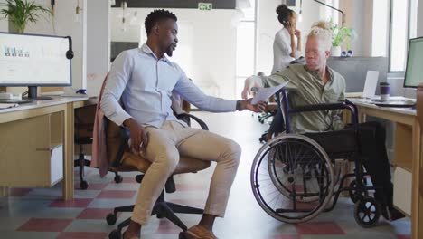 Diverse-business-people-discussing-with-disabled-colleague-and-documents-in-creative-office