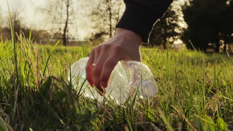 recogiendo una botella de agua de plástico vacía tirada en el parque de césped, la mano femenina agarra basura, hora dorada, cierre estático