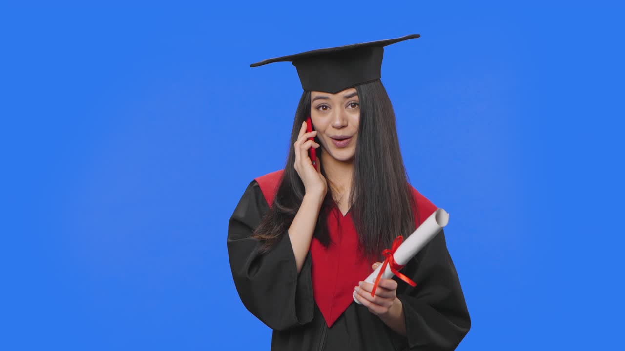 Portrait of female student in graduation costume, holding diploma and talking on a smartphone. Young brunette woman posing in studio with blue screen background. Close up. Slow motion ready 59.94fps