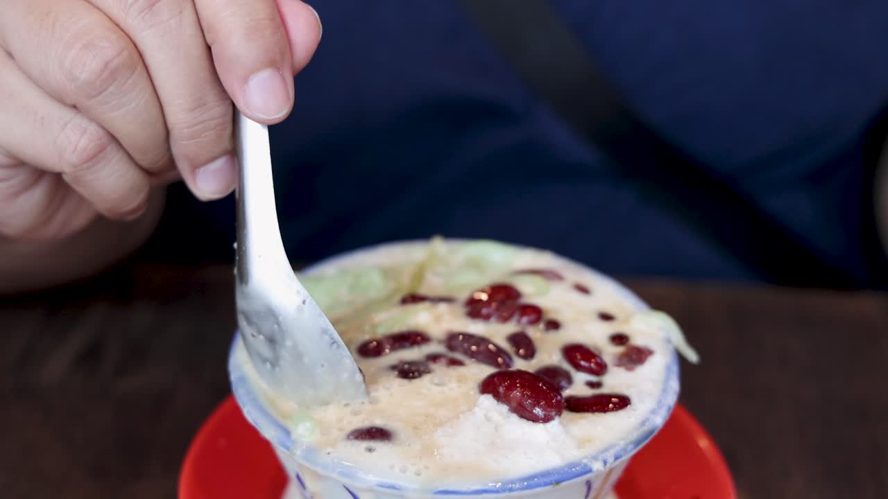 Close-up of hand scooping cendol dessert with red beans in a cozy indoor setting