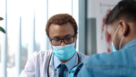 Young-Handsome-Male-Physician-Specialist-In-Medical-Mask-Talking-With-Man-Patient-Explaining-Coronavirus-Vaccine-At-Doctor-Consultation,-Covid-19-Virus-Cure,-Vaccination-Concept