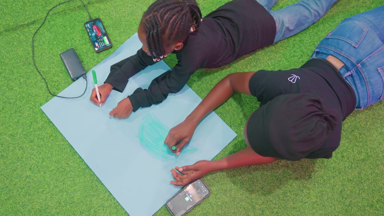 Black mother and child creating outdoors on grass with blue sheet, collaborative doodling and coloring, mother as creative partner, playful gestures, phone and charger visible, casual jeans, braided