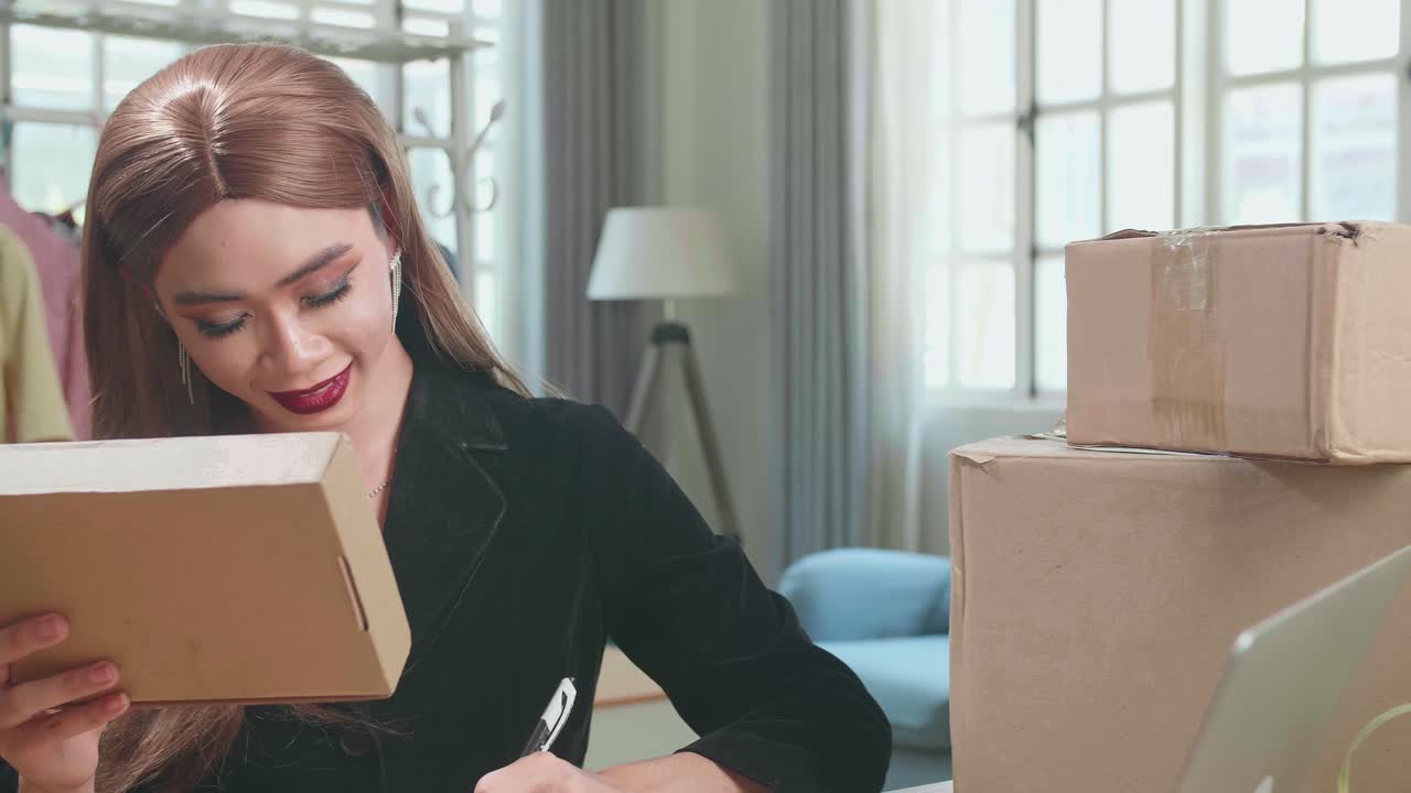 Young Asian Transgender Woman Business Owner With Boxes On The Table Happy Online Sales Job, Use The Laptop, Get An Order From Customers, Take Notes, And Make Arrangements For Delivery By Post