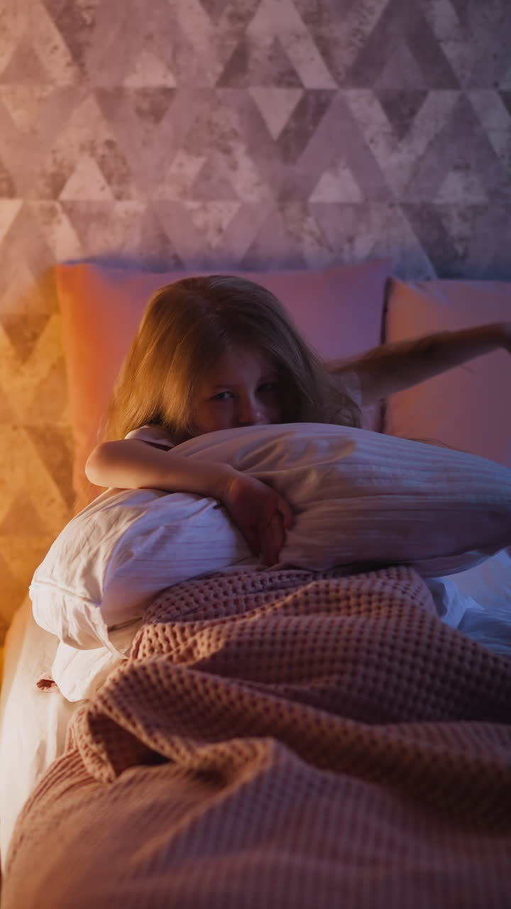 Distressed little girl hugs soft pillow and breathes heavily sitting on bed with knitted plaid near illuminated play teepee in bedroom at night slow motion