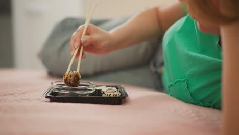 woman with polished nails lying on bed dips sushi roll into small sauce bowl using chopsticks lifting roll slowly to let excess sauce drip off calm casual setting with green shirt and pink blanket