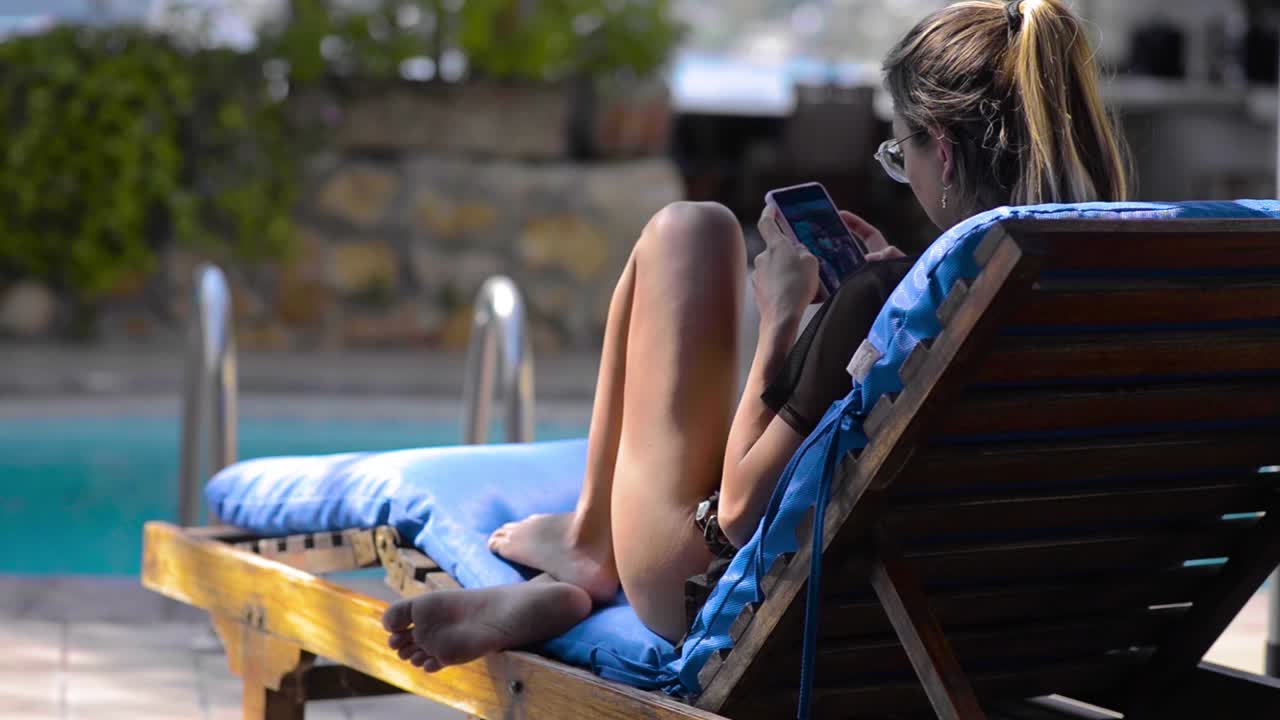 Young latin girl lying on a sunbed on a warm sunny day using her mobile near the pool