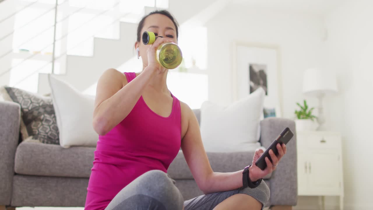 Donna asiatica concentrata sul tappetino, che riposa, beve e usa lo smartphone.