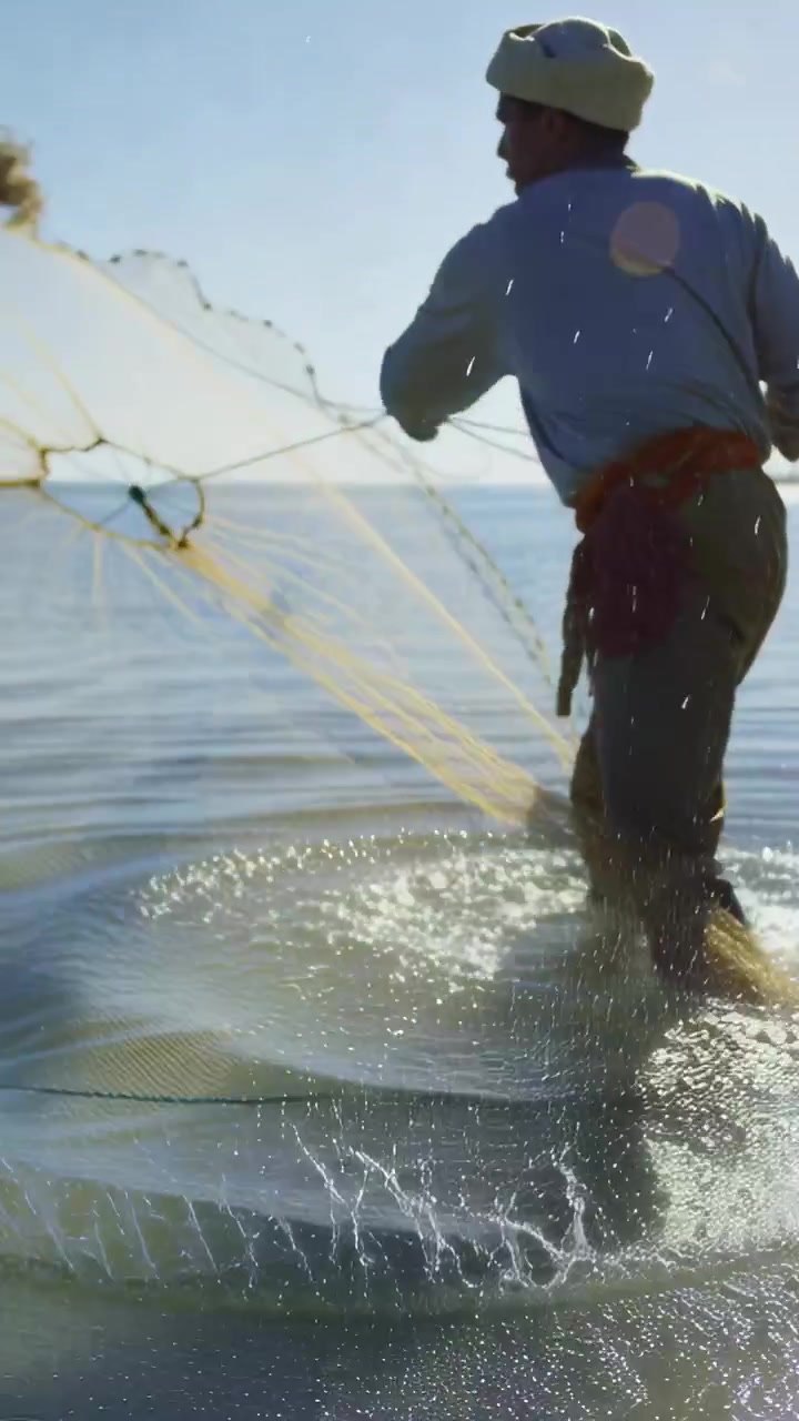 Video verticale: Uomo con turbante che cammina e lancia una rete da pesca in mare, catturando pesci, con spazio per il testo.