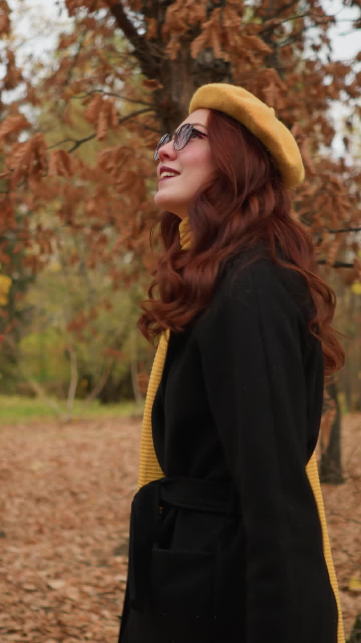 Sidebilde av dame som spaserer gjennom skogen, iført gult skjerf, solbriller, beret og svart kåpe, omgitt av høstløv, nyter naturen med et rolig og avslappet uttrykk.
