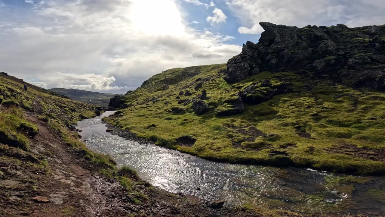 Sakte film-opptak av en klar bekk som renner gjennom en smal vulkansk steinkløft på Island, dekket av frodig grønn mose. Kameraet panorerer for å avsløre den ulendte kløften med en tursti av brun jord.