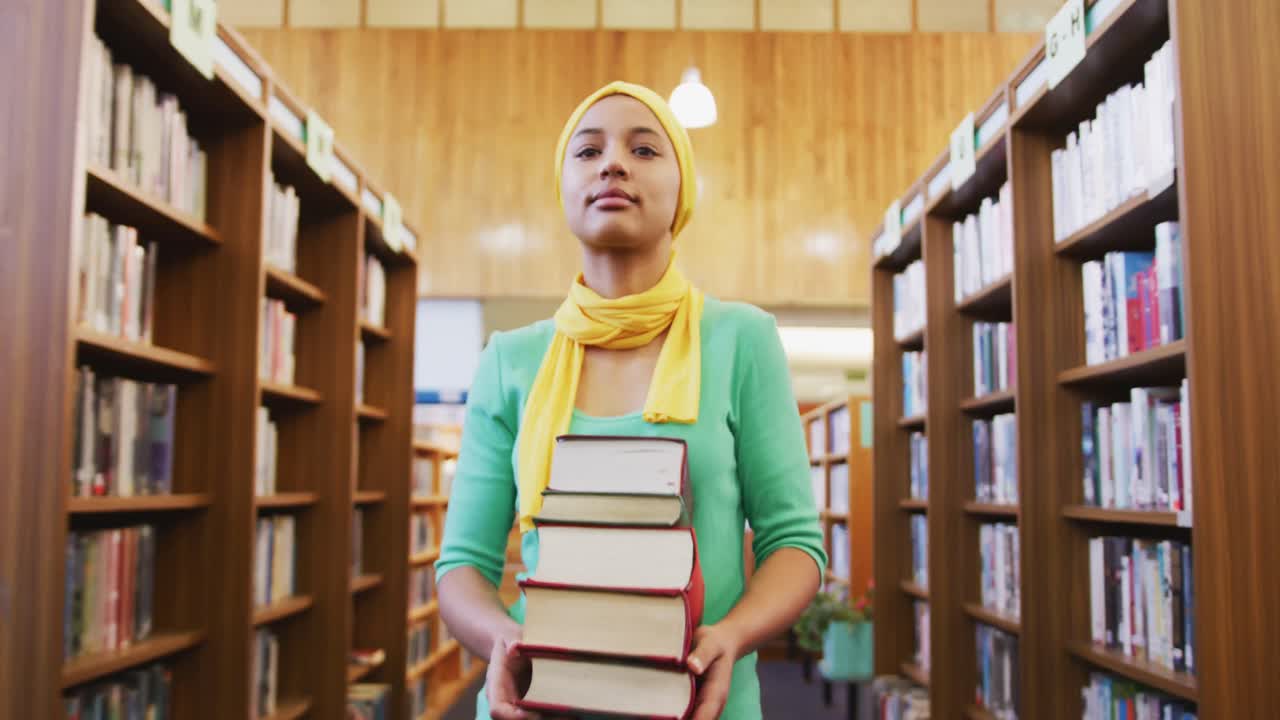 una estudiante asiática con un hijab amarillo caminando entre estantes de libros y sosteniendo una pila de boo