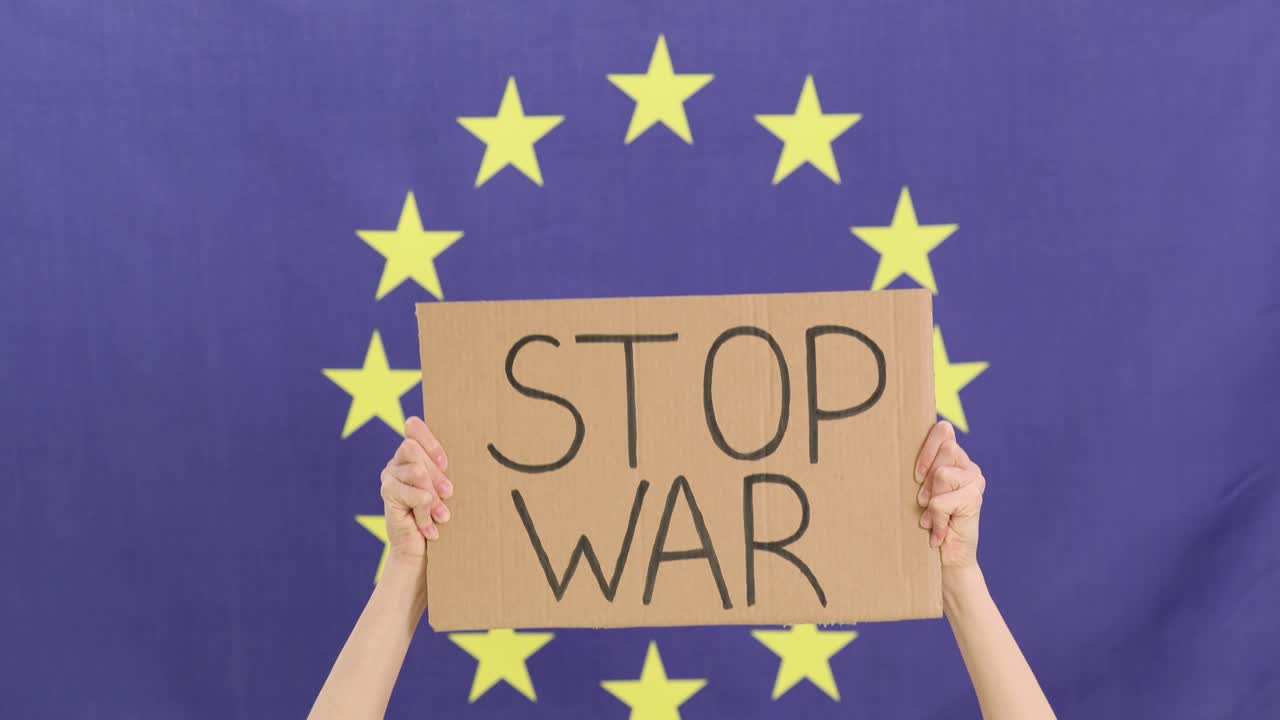 Mani sollevano un cartello "stop alla guerra" contro la bandiera dell'Unione Europea.