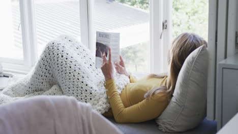 Mujer-Birracial-Sentada-Junto-A-La-Ventana-Leyendo-Un-Libro-En-Casa,-Cámara-Lenta