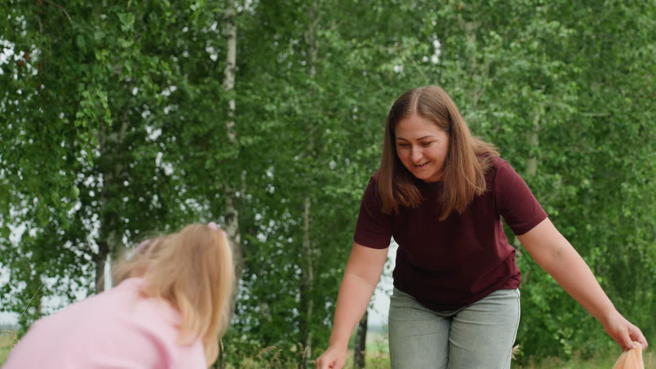 White mother and child with blanket smiling woman unfolds orange blanket while little girl watches, birch trees and green meadow backdrop, warm maternal interaction, playful anticipation and gentle