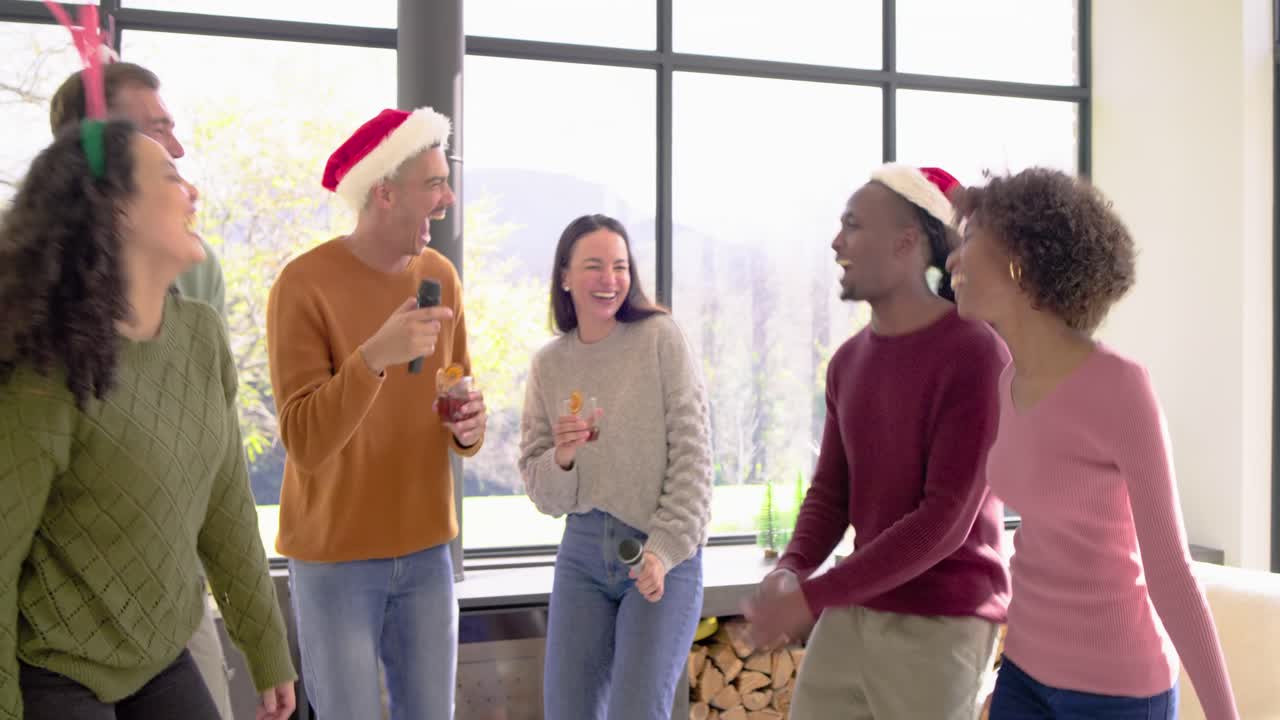 Diverse friends lifting smartphone above group at home, singing karaoke by stove for holiday party