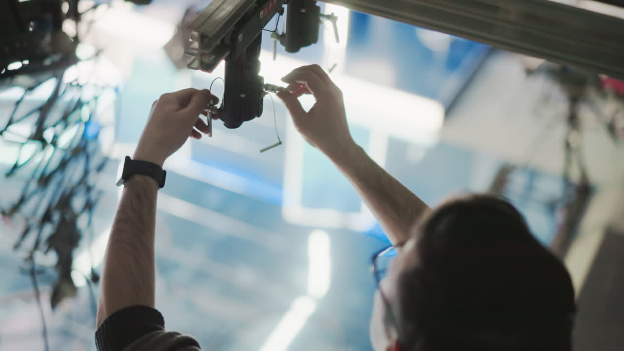 Technician handling lighting rigging system, adjusting control unit with tools, focusing on fine adjustments in professional studio setting, preparing for production setup, ensuring precise alignment