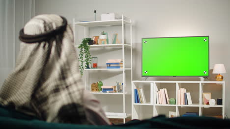 middle eastern man watching tv with chroma key close-up. person watches television with green screen in living room. wearing traditional islamic clothes