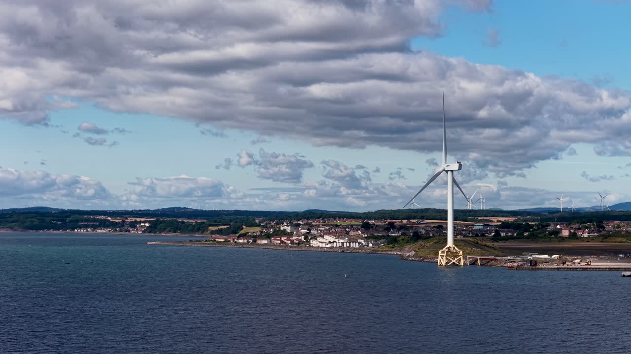 Large offshore wind turbine spins steadily beside coastal town under partly cloudy daylight sky