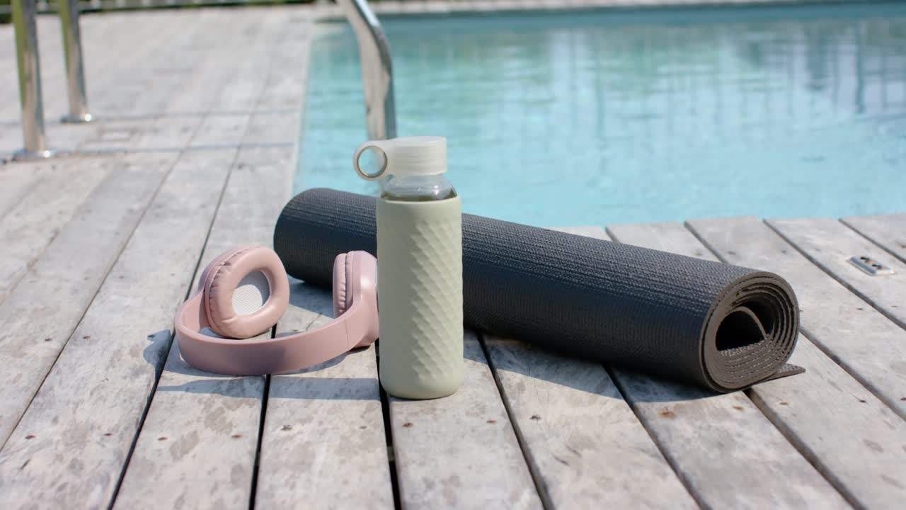 Tappetino da yoga grigio, borraccia verde e cuffie rosa su una piattaforma a bordo piscina, pronti per un allenamento in piscina