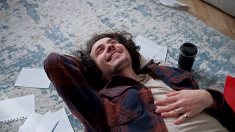 happy brunette guy with curly hair in a checkered shirt lies on a pillow on the floor surrounded by sheets of paper and a cup of coffee at home while working