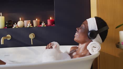 Smiling-african-american-woman-with-towel-taking-bath-and-wearing-headphones-in-bathroom