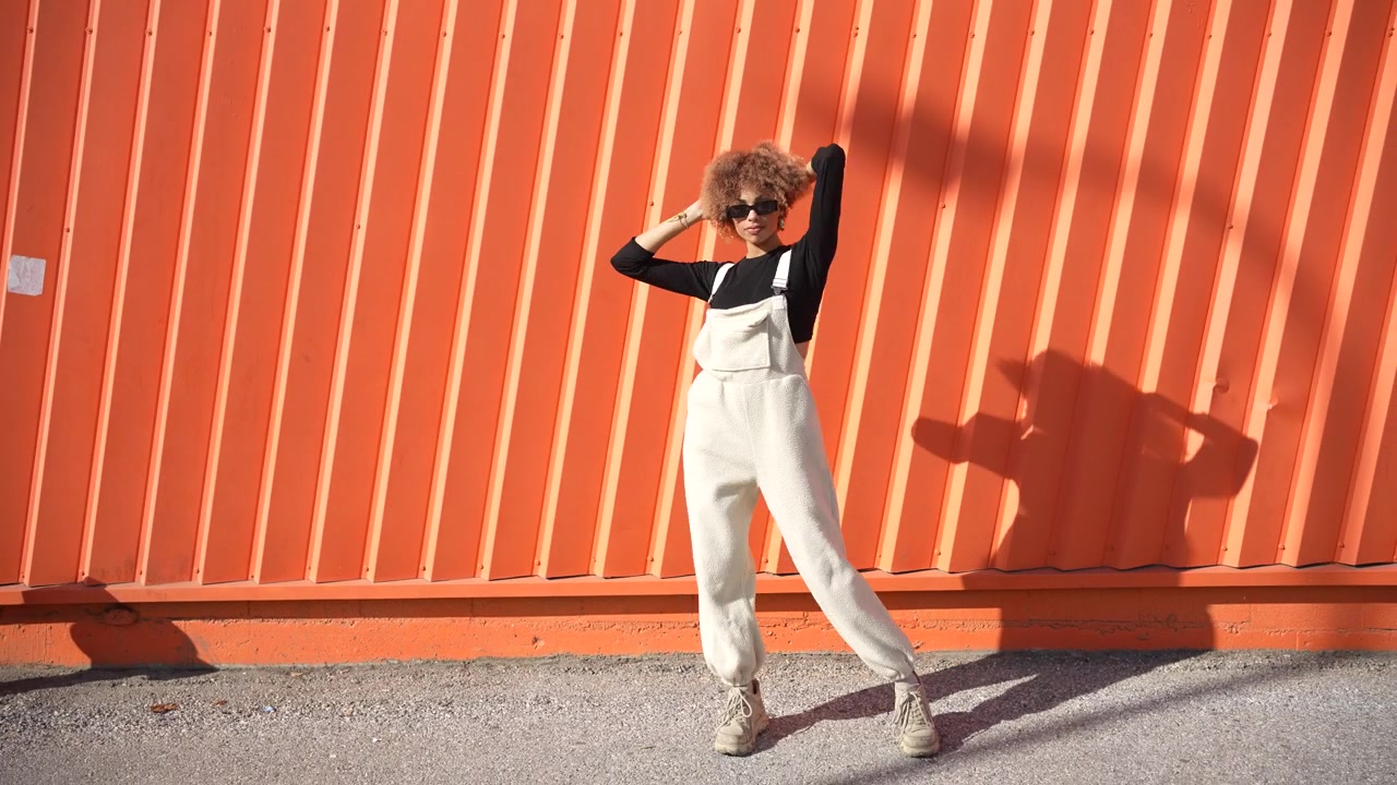 Eine stilvolle Frau posiert selbstbewusst vor einer leuchtend orangefarbenen Wand, wunderschön festgehalten.