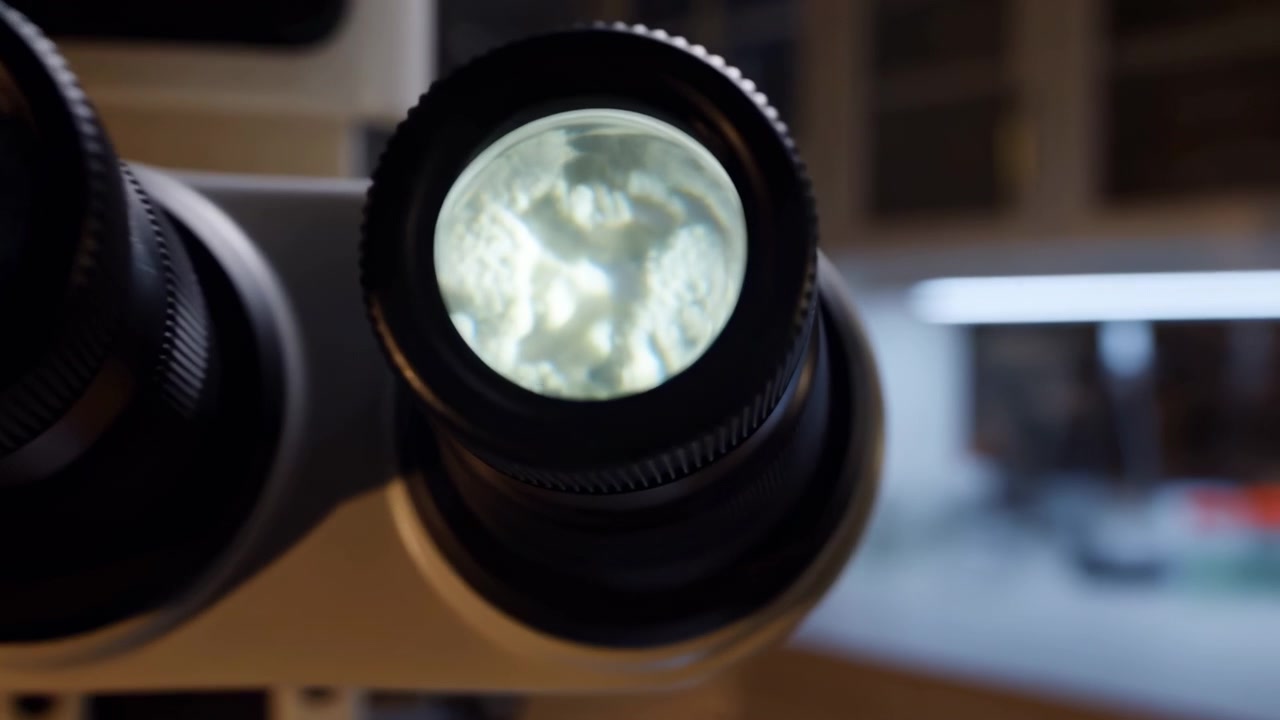 Una vista ravvicinata di un moderno microscopio binoculare poggiato su un banco scuro in un laboratorio, con armadietti illuminati delicatamente e un'area di lavoro sfocata sullo sfondo