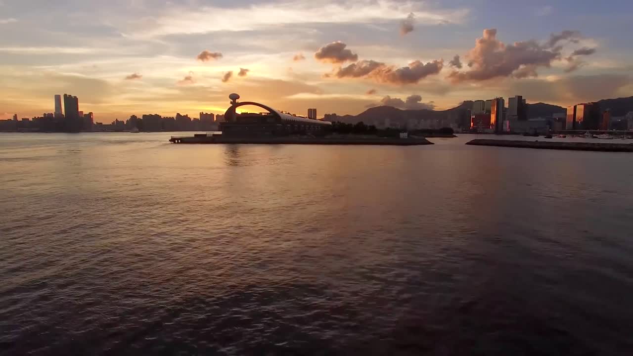 Aerial footage or a drone shot over a sea or water in a harbor while a city is visible in the background in silhouette mode while the sun is setting or sundown. Clouds are visible and sky is orange.