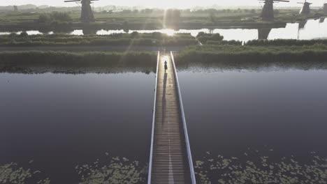 a drone shot panning down, while a girl walks on a bridge, with dutch windmills in the netherlands during sunrise