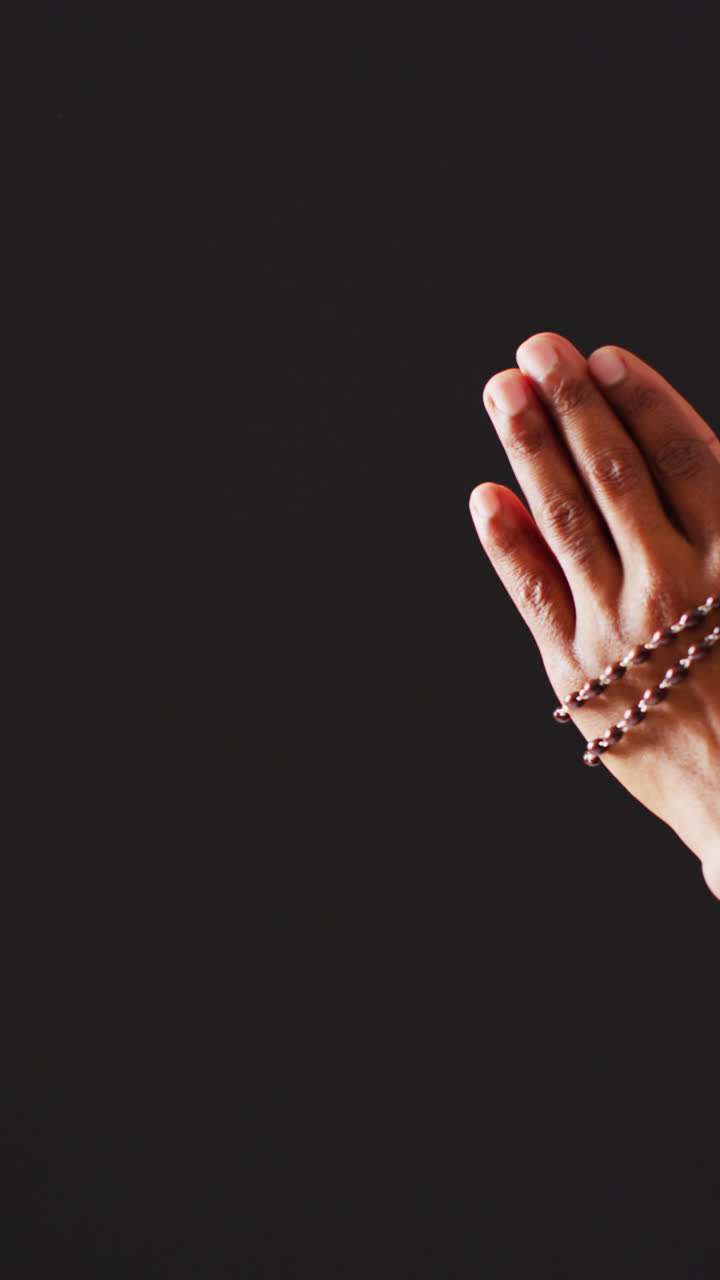 Vertical video of praying hands with rosary on black background with copy space