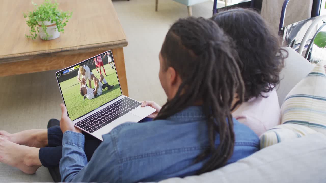 video de dos personas afroamericanas sentadas en el sofá y viendo un partido de fútbol en una computadora portátil