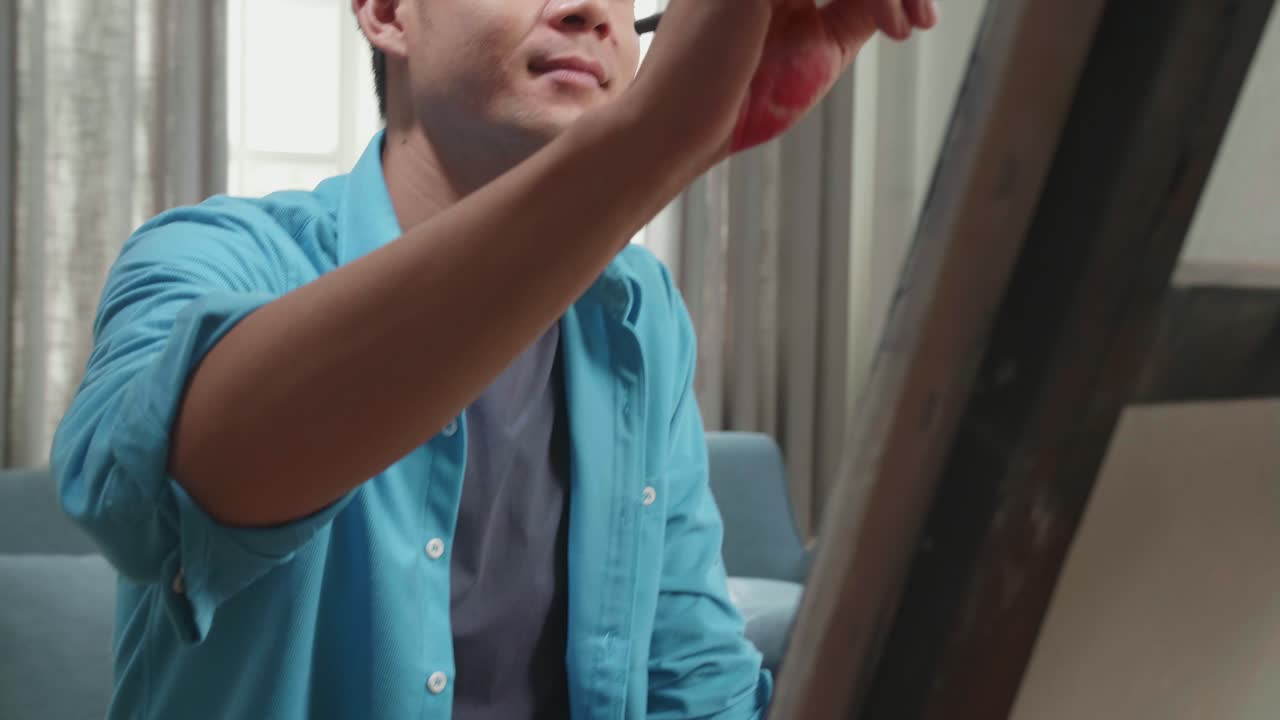 Close Up Of An Asian Artist Man Sitting On Round Wooden Chair Without Backrest Holding Paintbrush And Mixing It With Colour Before Painting On The Canvas