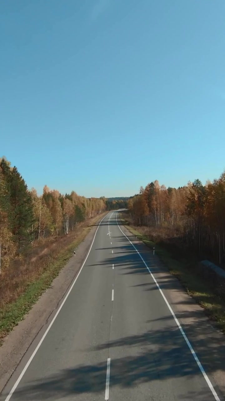Záběry z dronu opuštěné dvouproudové silnice táhnoucí se do dálky, obklopené barevnými podzimními stromy a jasnou modrou oblohou, téma cestování a dopravy.