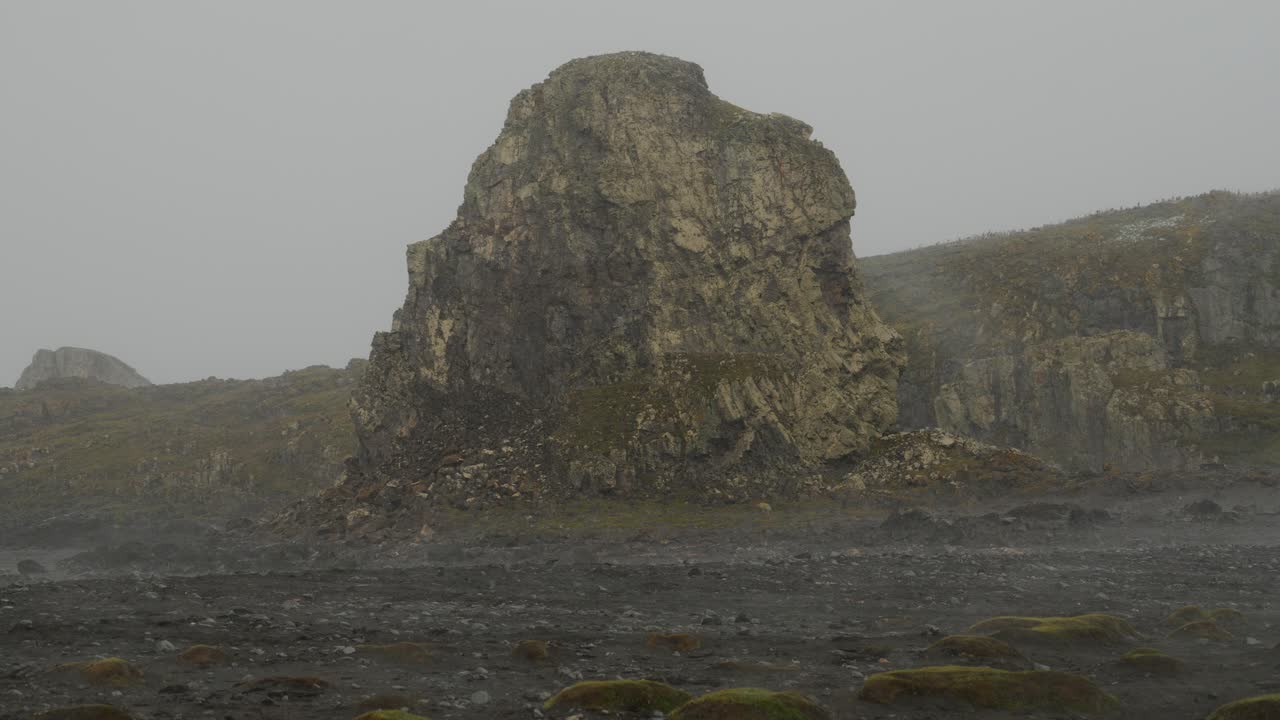 Massive jagged volcanic rock formation rises from dark moss-covered coastal plain at Point Wild, Elephant Island, Antarctica, during light snowfall under heavy grey overcast skies