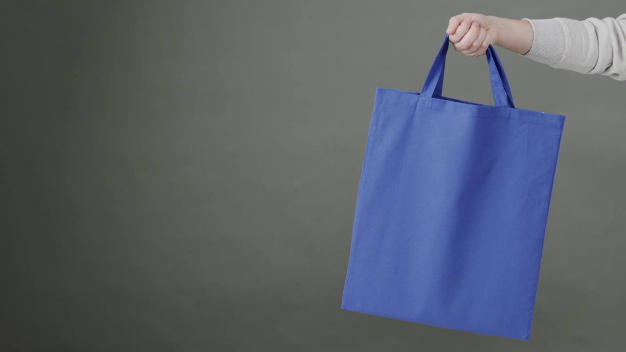 mano de mujer caucásica sosteniendo una bolsa azul sobre fondo gris, espacio de copia, cámara lenta