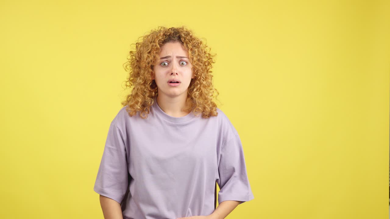 Young woman with curly hair reacting with shock and fear against a yellow background