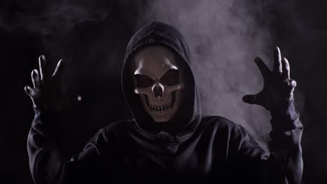 close up of scary man in the hooded sweatshirt wearing halloween mask making frightening gesture on the black background with smoke