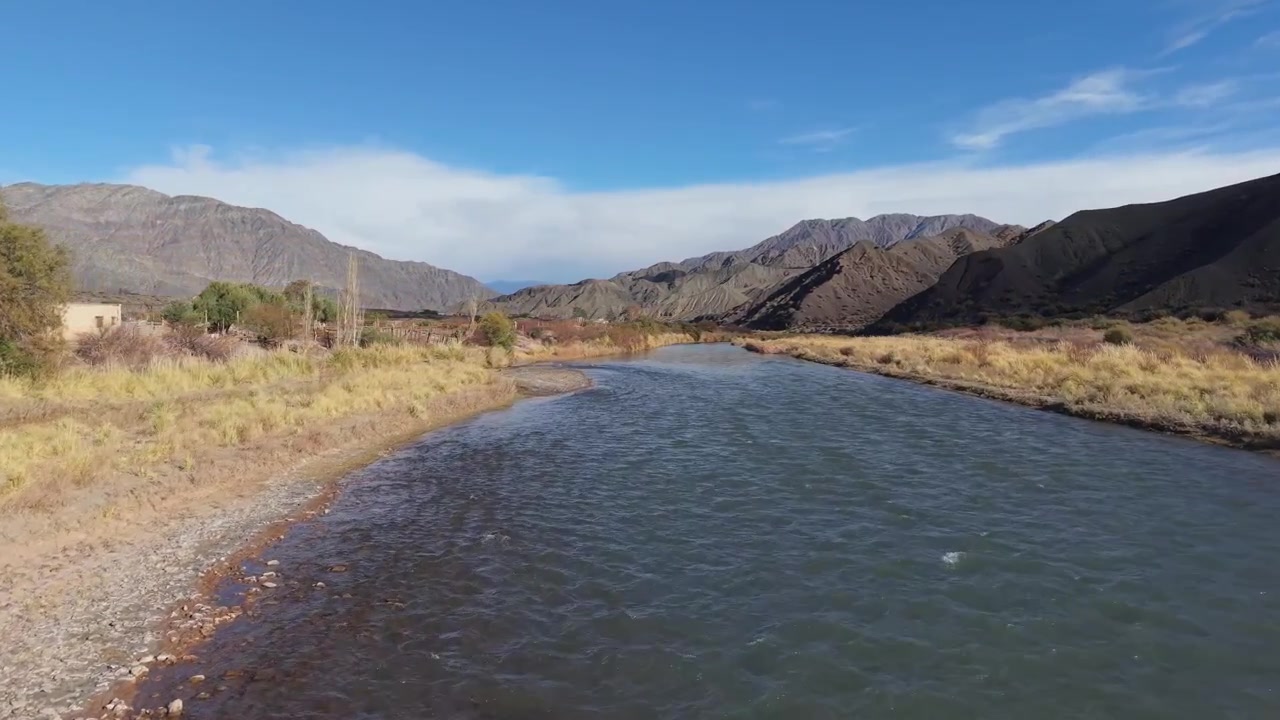 Tomada suave de drone desliza acima do Rio San Juan, capturando margens sinuosas, grama dourada árida e montanhas andinas dramáticas sob luz do dia clara e brilhante.