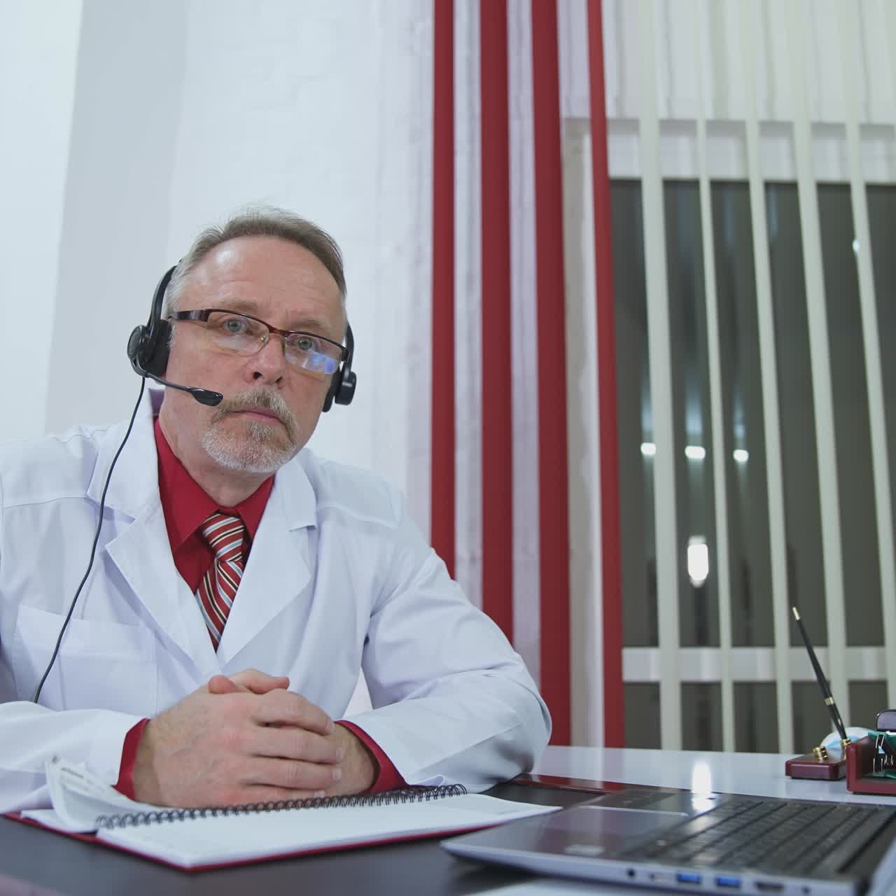 Medico anziano ascolta il paziente durante una videochiamata. Anziano medico in uniforme e con cuffie che effettua una consultazione a distanza. Concetto di telemedicina.