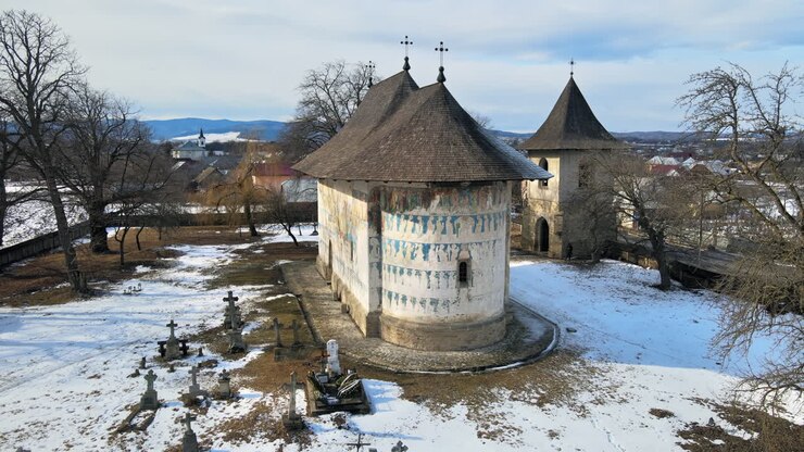 Aerial drone view of The Arbore Monastery in winter, Romania. Main painted church, yard with snow and cemetery, bare trees, residential buildings