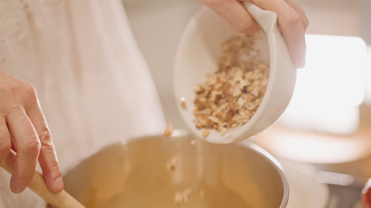 Versando noci pecan tritate nel ripieno caldo, cucchiaio di legno che guida le noci in una ciotola di metallo, mani in primo piano, preparazione del topping croccante, luce naturale, scena di cucina rustica, pasticcere che finisce lo strato della torta di zucca