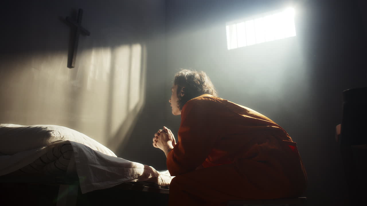 Inmate Kneeling beside Bed and Praying to God in Prison Cell