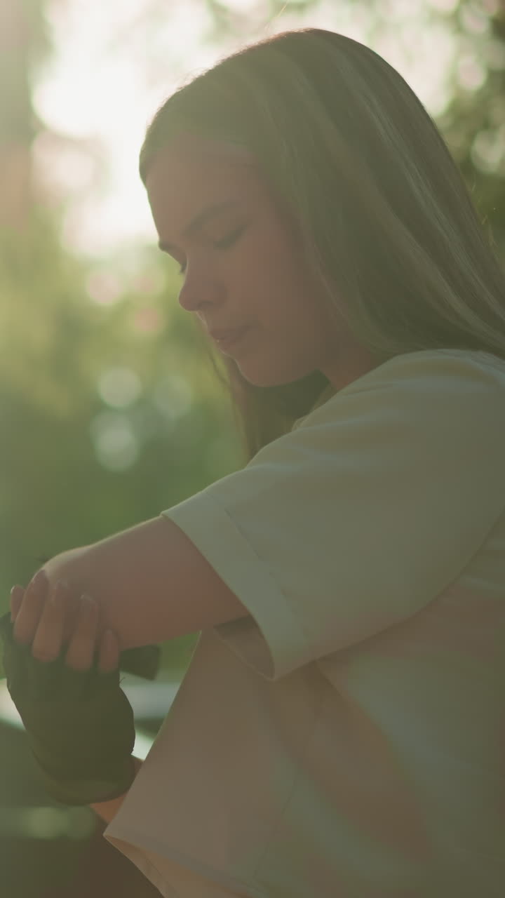 검은 장갑을 입은 젊은 성인은 팔꿈치를 부드럽게 문지르며, 빛은 울창한 초록색 잎을 통해 필터링되며, 그녀에게 따뜻한 빛을 며, 지나가는 차와 함께 흐릿한 배경입니다.